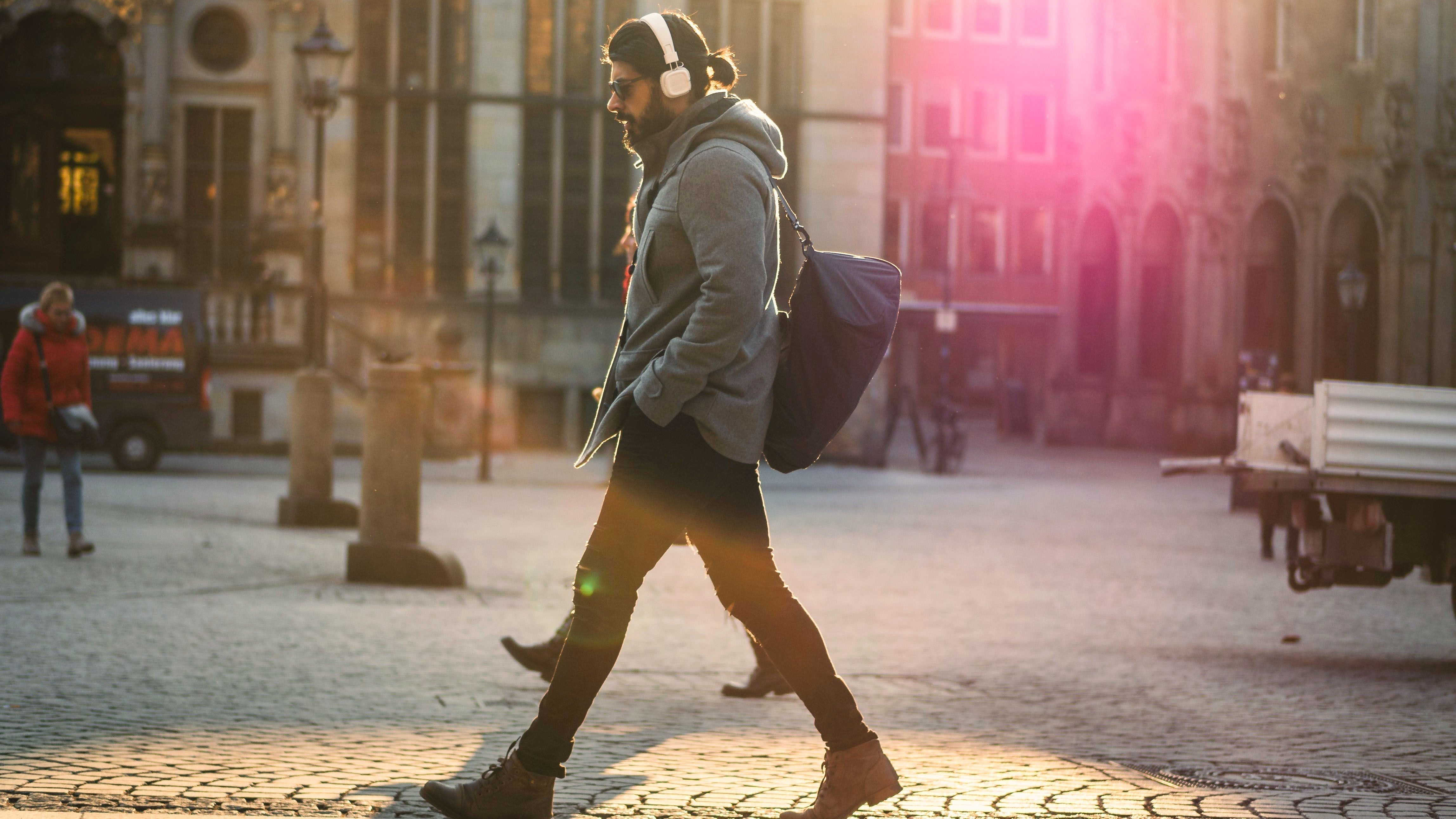 Mann mit weißen Over-Ear-Kopfhörern geht durch sonnige Stadt – Vergleich kabelgebunden vs. kabellos: Welche sind sicherer?
