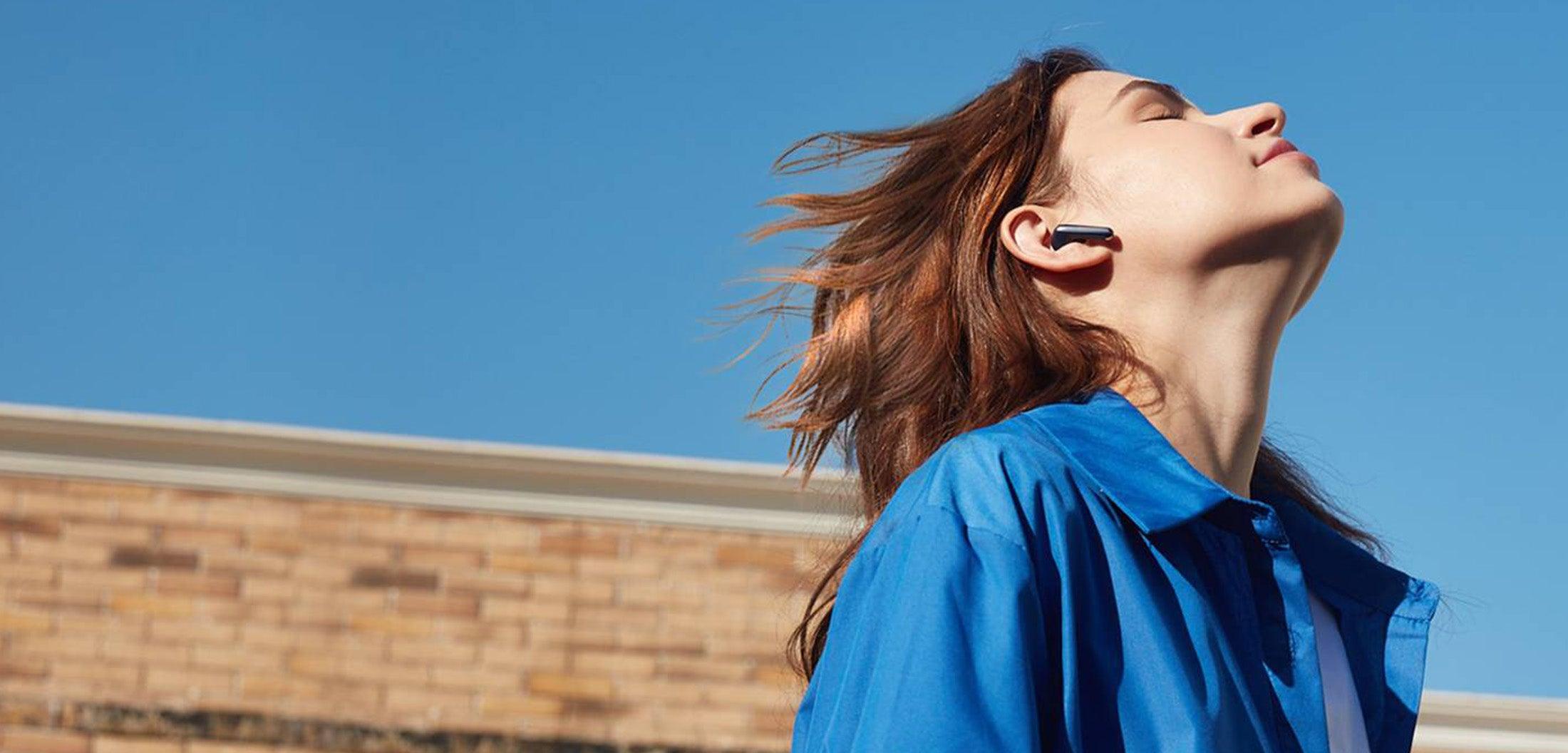 Frau mit Bluetooth-Ohrhörer genießt Musik unter blauem Himmel – für Audiophile, die überlegene Klangqualität suchen.