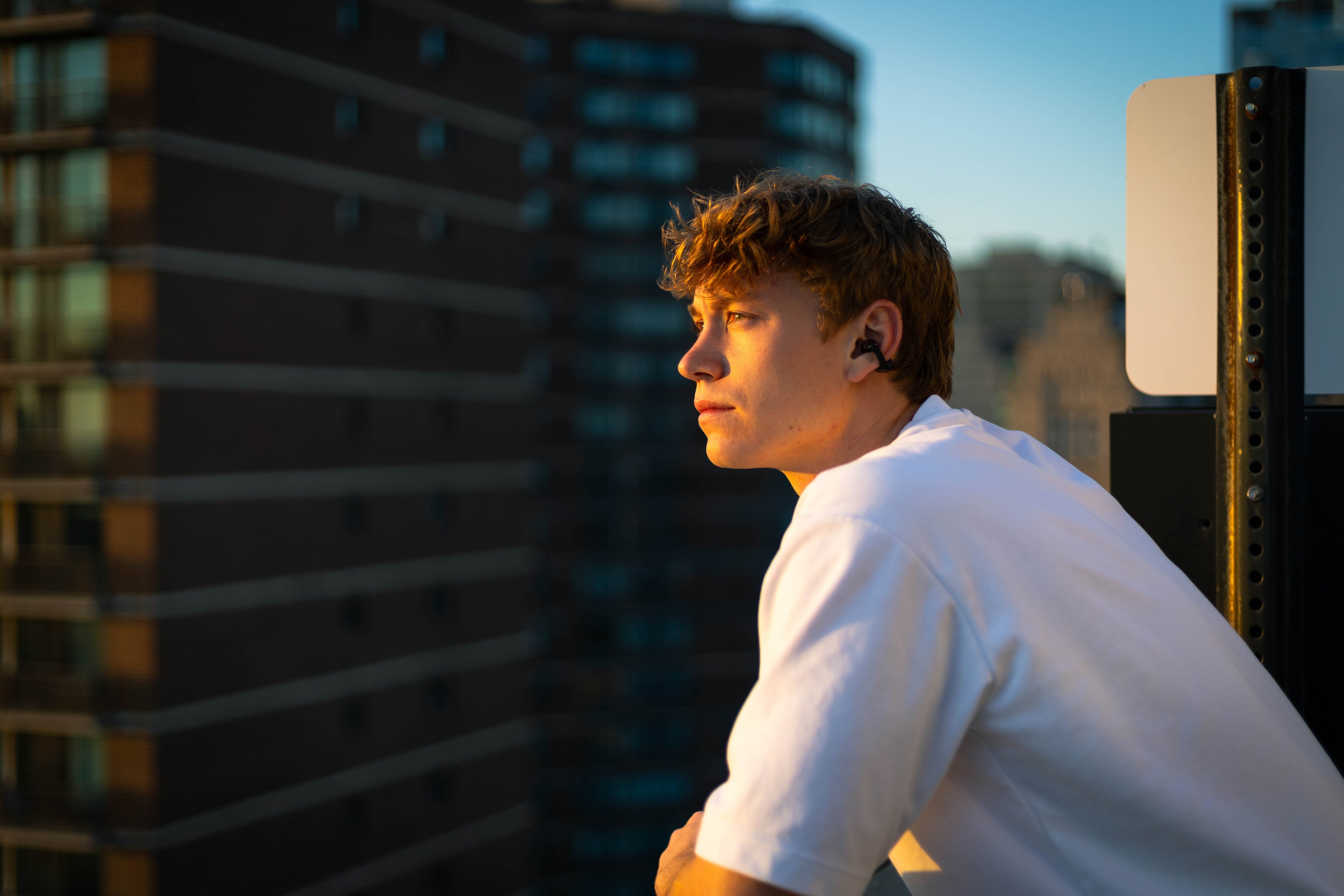 Junger Mann mit Bluetooth-Kopfhörern auf dem Dach bei Sonnenuntergang.