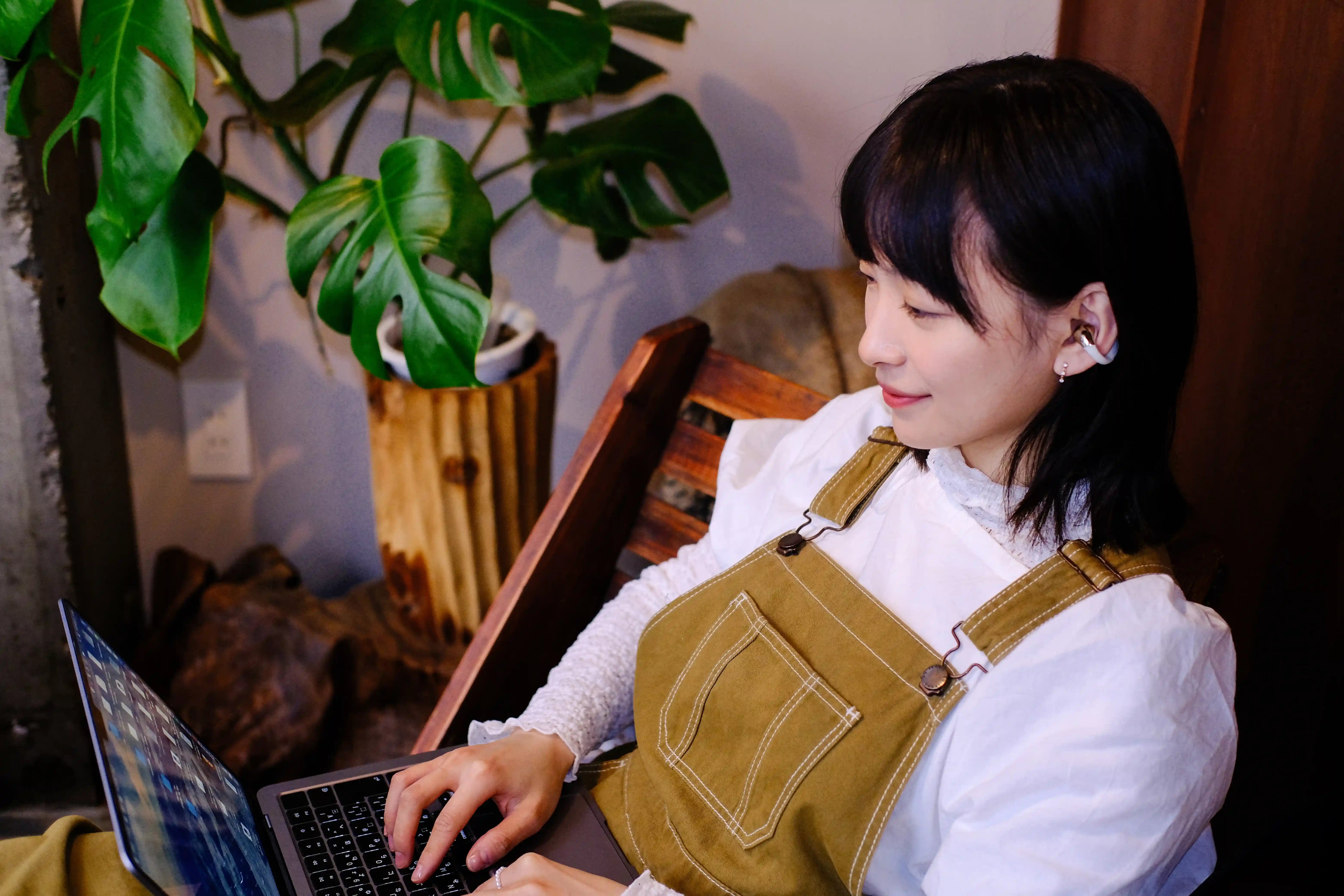 Frau mit Open-Ear-Kopfhörern arbeitet entspannt am Laptop in gemütlichem Raum mit Zimmerpflanze im Hintergrund.