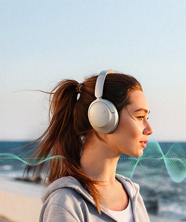 Mulher com auscultadores over-ear brancos na praia, o vento sopra no cabelo.