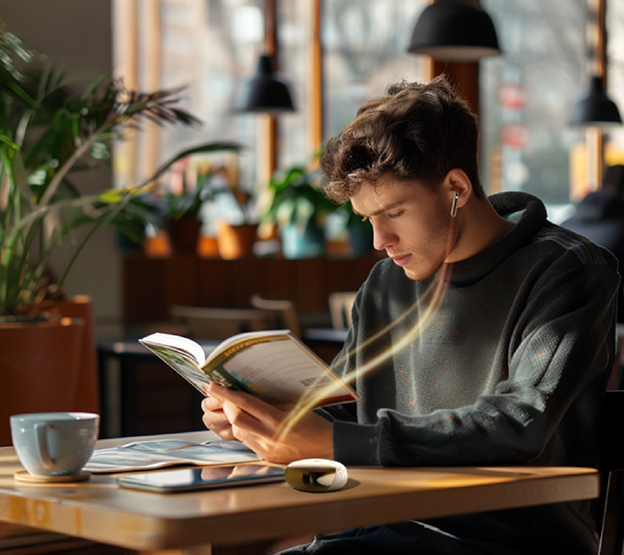 Jovem homem lê um livro num café com auscultadores sem fios e uma chávena de café sobre a mesa de madeira.
