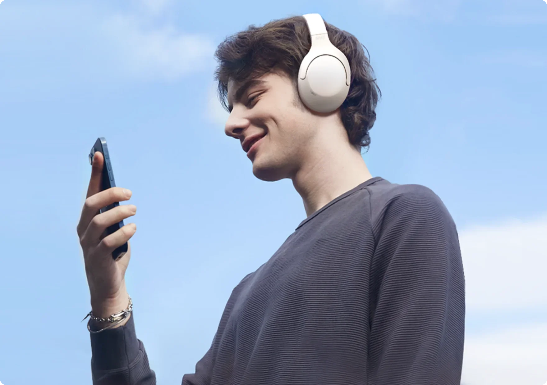 Jovem com auscultadores brancos e smartphone sob um céu azul.