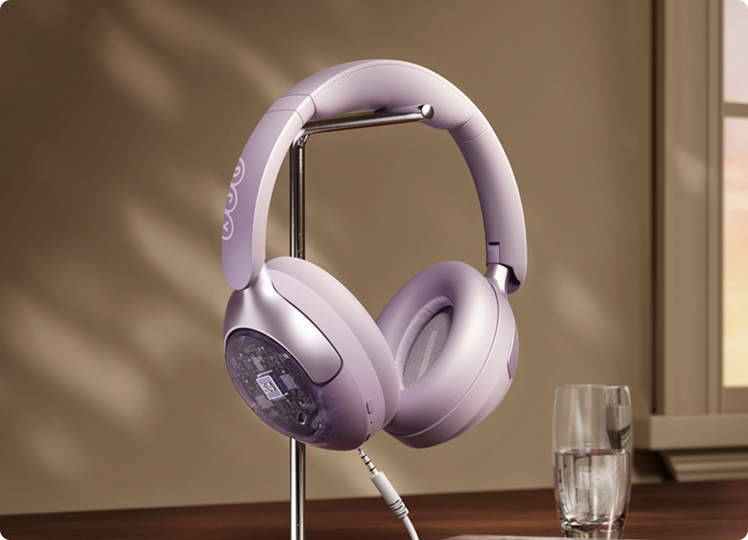 Auriculares over-ear cor de lavanda com caixa transparente num suporte, cabo ligado, ao lado de um copo de água na janela.